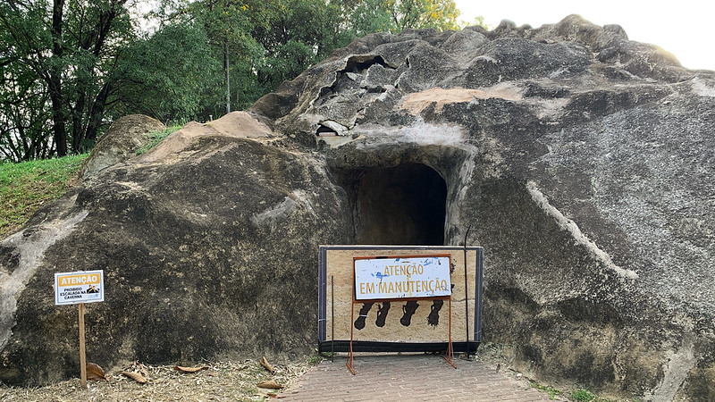 A imagem mostra uma réplica de uma grande formação rochosa com uma abertura semelhante a uma caverna no centro. Na entrada, há uma placa dizendo Atenção: Em manutenção, com ilustrações de pegadas. Um cercado de madeira e metal bloqueia o acesso à caverna. À esquerda, outra placa avisa sobre possíveis desmoronamentos. O local está rodeado por árvores e vegetação.