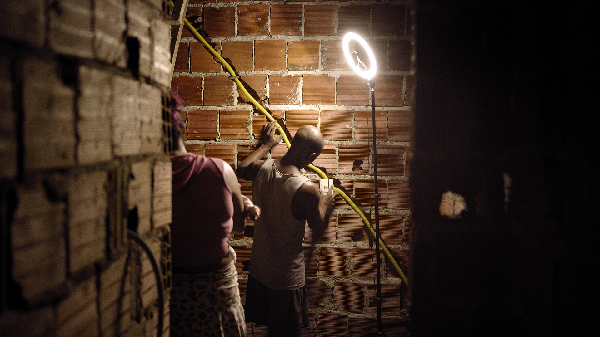 A imagem mostra um ambiente de construção simples, com paredes de tijolos expostos sem reboco. Em primeiro plano, duas pessoas estão de costas para a câmera. A pessoa à direita é um homem negro, de cabeça raspada, que parece estar concentrado em um trabalho na parede. Ele segura um fio amarelo que atravessa a parede em diagonal, enquanto parece manusear uma tomada ou interruptor. A outra pessoa, à esquerda, é uma mulher, também de costas, vestindo uma camiseta rosa e um shorts com estampa. No canto superior direito, há um anel de luz em um tripé, iluminando a cena de forma suave, contrastando com a penumbra ao redor. A cena sugere um trabalho de instalação elétrica em andamento, em um ambiente residencial ou de construção.