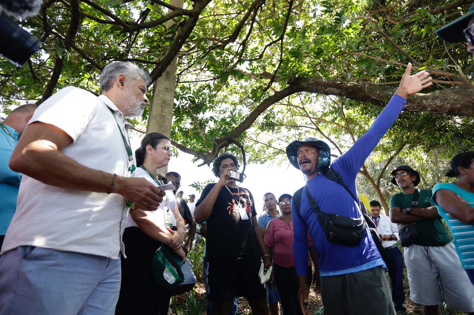 A imagem mostra um grupo de pessoas reunidas ao ar livre, sob a sombra de árvores. Há várias pessoas na foto, algumas segurando papéis ou cadernos, e um homem negro de bigode, vestindo uma camisa azul de mangas longas, à direita está com o braço levantado, apontando para algo fora da imagem. À esquerda, um homem branco, de barba e cabelos brancos, acompanha a fala do homem negro. Algumas pessoas estão usando crachás ou identificações penduradas no pescoço. A vegetação ao redor é densa, com muitas folhas e galhos. A luz do sol penetra pelas folhas, criando um ambiente iluminado e natural.