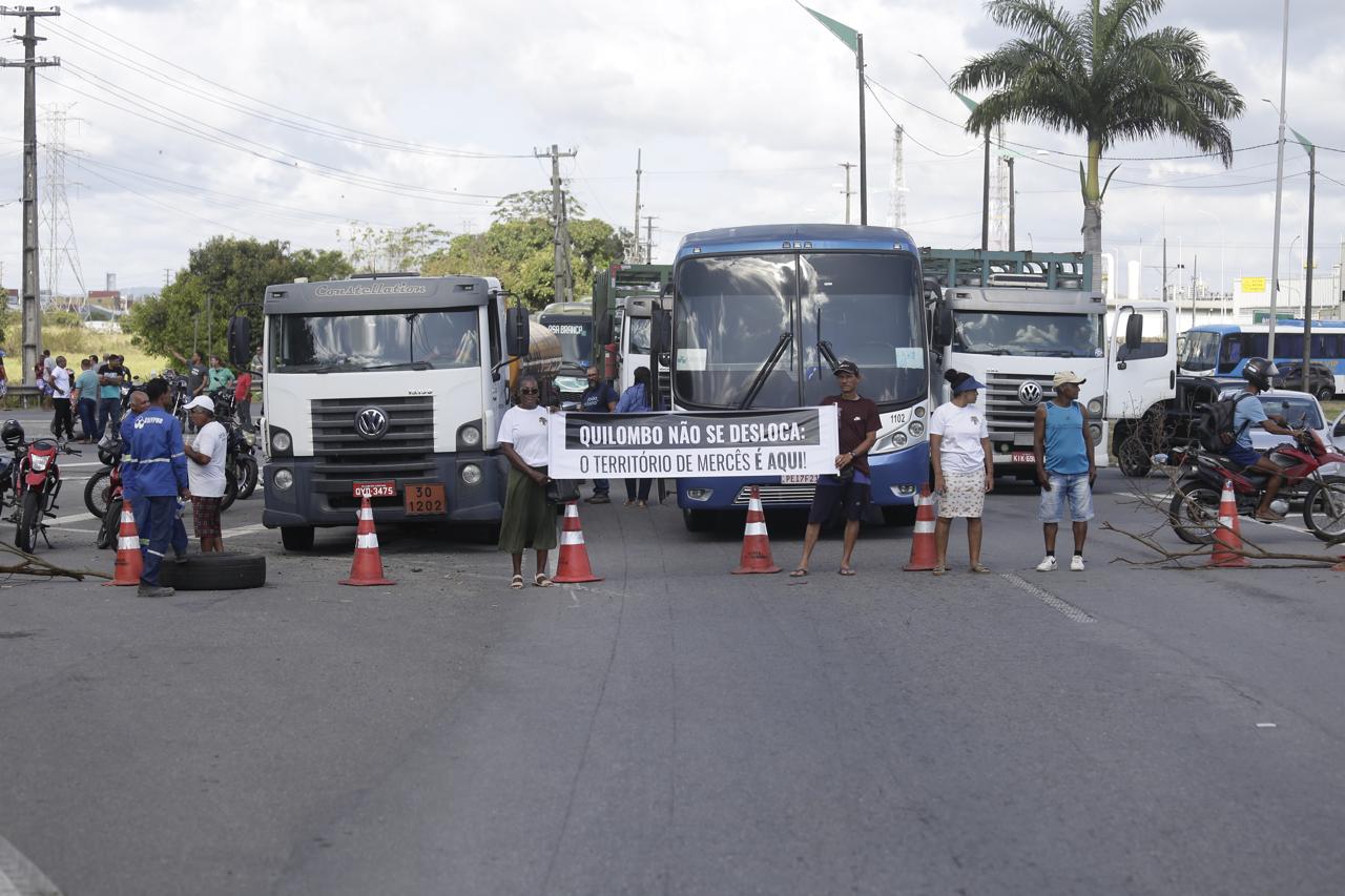 A imagem mostra uma manifestação em uma estrada, onde várias pessoas estão bloqueando a passagem de veículos, incluindo caminhões e ônibus. Os manifestantes seguram um grande cartaz que diz: QUILOMBO NÃO SE DESLOCA: O TERRITÓRIO DE MERCÊS É AQUI!. Há cones de trânsito laranja e branco posicionados na estrada para ajudar a bloquear o tráfego. Algumas pessoas estão em pé ao lado de motocicletas, e há outras pessoas ao fundo, aparentemente participando ou observando a manifestação. A manifestação parece estar ocorrendo em uma área urbana, com postes de eletricidade e vegetação ao fundo.