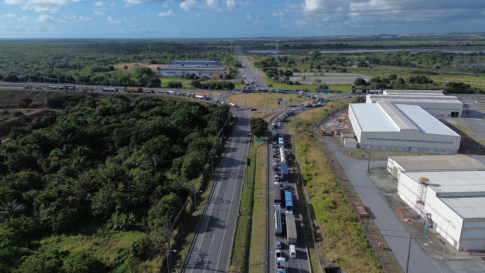 A imagem mostra uma vista aérea de uma estrada movimentada, com veículos parados em ambos os sentidos. A estrada principal se divide em duas direções, com uma rotatória no centro. À esquerda da estrada, há uma área densamente arborizada com vegetação verde. À direita, há vários edifícios industriais ou armazéns com telhados brancos. A área ao redor da estrada e dos edifícios é predominantemente verde, com algumas áreas abertas e campos ao fundo. O céu está parcialmente nublado, com algumas nuvens brancas, e o horizonte é visível ao longe. A imagem destaca o contraste entre a área industrial e a vegetação natural, além de mostrar o tráfego intenso na estrada.