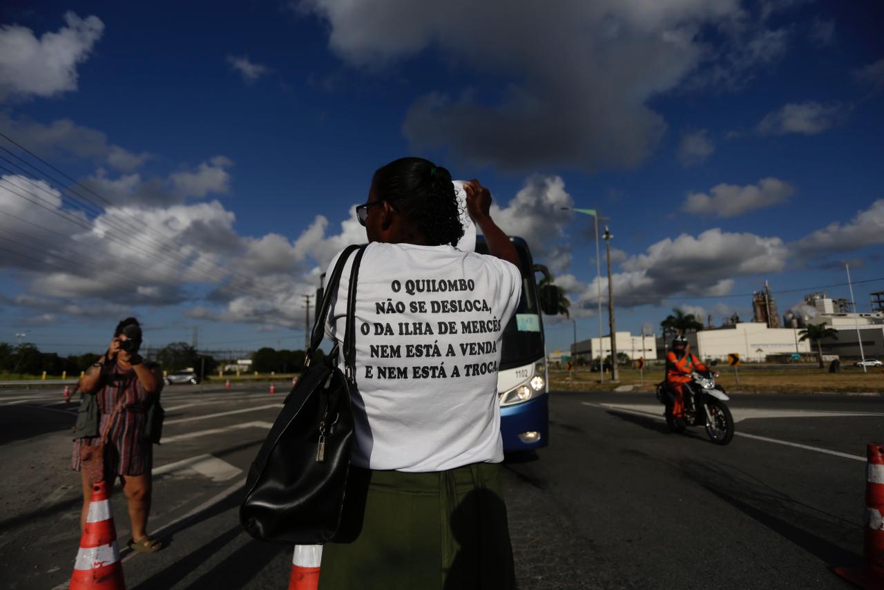 Você Imagem carregada descrever essa foto para uma pessoa cega Copilot A imagem mostra uma pessoa de costas, vestindo uma camiseta branca com a frase: O QUILOMBO NÃO SE DESLOCA, O DA ILHA DE MERCÊS NEM ESTÁ À VENDA E NEM ESTÁ À TROCA. A pessoa está em uma rua, aparentemente participando de uma manifestação ou protesto. Ao fundo, há outra pessoa tirando uma foto e um motociclista passando. O céu está parcialmente nublado e há alguns prédios e palmeiras ao fundo.