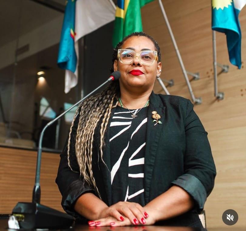 Foto de Elaine Cristina, vereadora do PSOL no Recife. Ela é uma mulher negra, de cabelos longos com tranças, usando óculos e vestindo um conjunto de casaco preto sobre blusa preta com grafismos brancos. Ela foi fotografada em uma tribuna com microfone. Ao fundo, há mastros de bandeiras das quais se vê apenas a parte inferior.