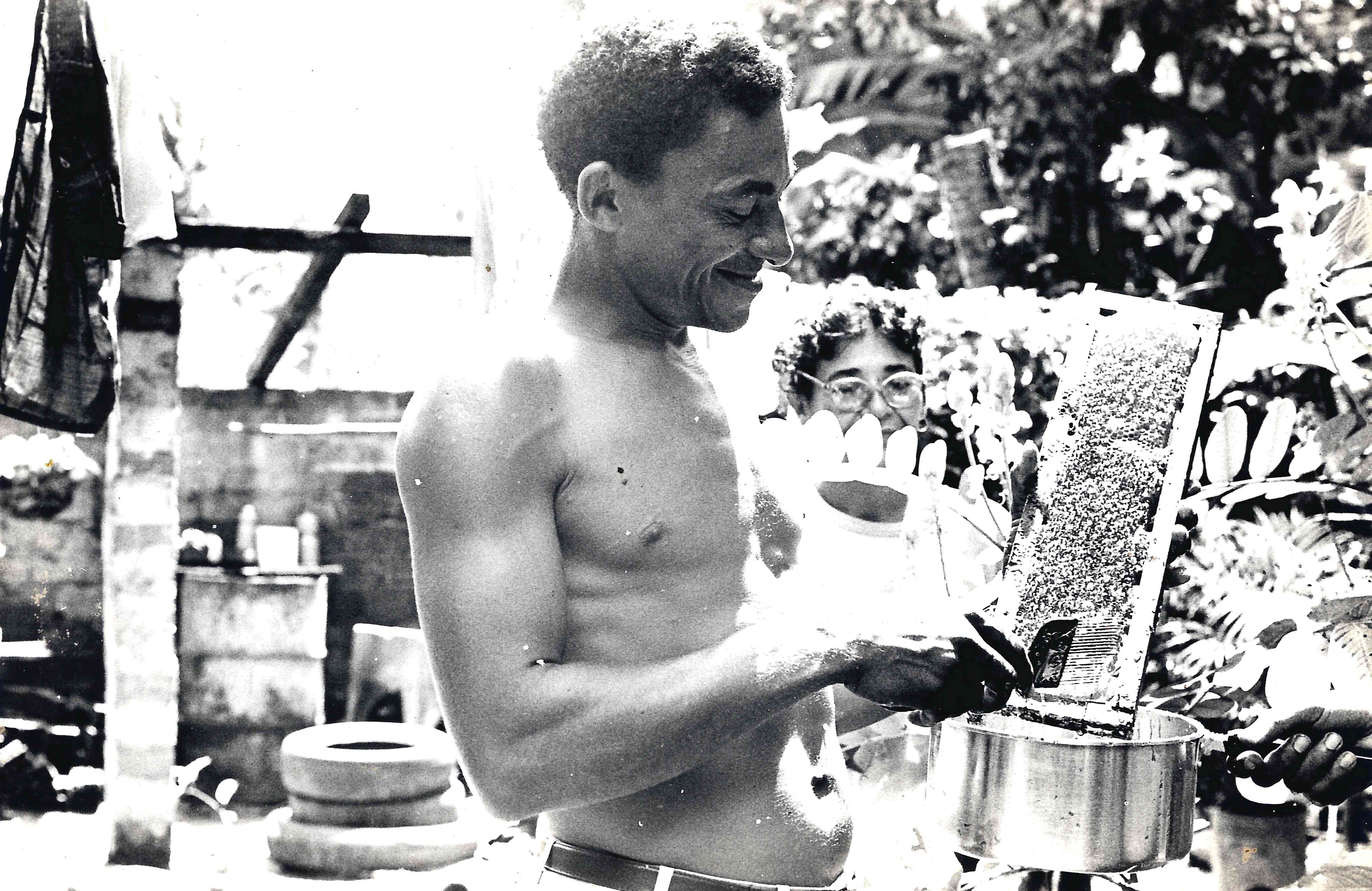 Uma fotografia em preto e branco que mostra um homem sem camisa, sorrindo, enquanto segura uma colmeia de abelhas. Ao seu lado, parcialmente em segundo plano, está uma mulher que observa a colmeia. Ambos parecem estar em um ambiente externo, com plantas ao fundo e um cenário rústico. O homem está com um utensílio nas mãos, aparentemente utilizado para coletar mel de abelhas.