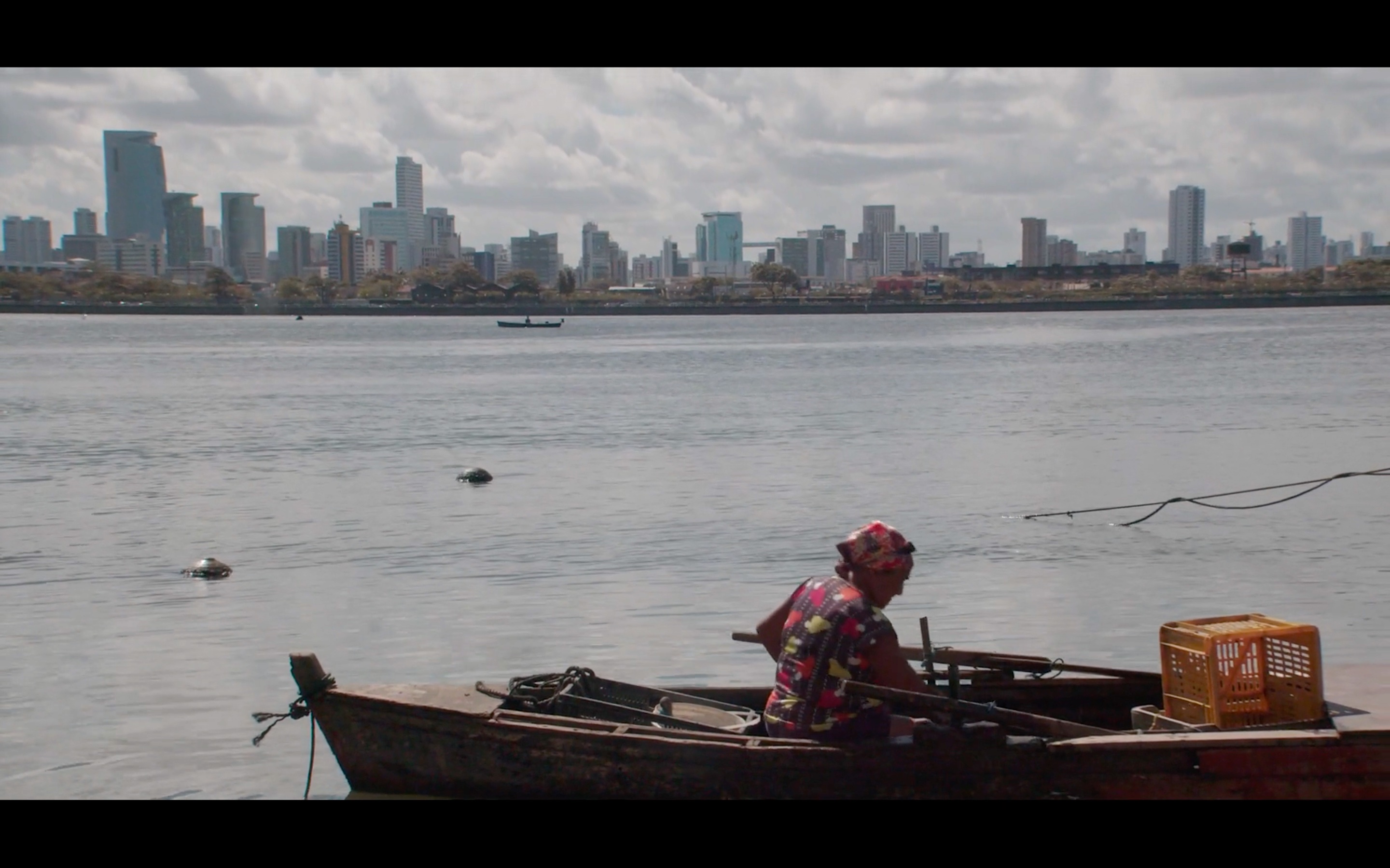 A imagem mostra uma pessoa em um barco pequeno, remando ou trabalhando, com uma cidade ao fundo que exibe prédios altos e modernos à beira de um corpo d'água. O céu está parcialmente nublado e a cena passa uma sensação de contraste entre o cenário urbano e a vida simples da pessoa no barco.