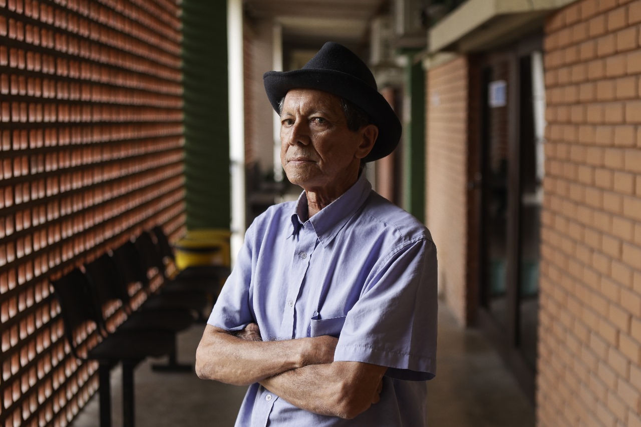 A imagem mostra Severino Vitorino, homem branco de pé em um corredor. Ele está usando uma camisa de manga curta de cor clara e um chapéu escuro. Os braços estão cruzados. O corredor tem paredes de tijolos à vista e há cadeiras pretas alinhadas ao longo da parede à esquerda. No fundo, há uma porta de vidro e uma parede verde. A iluminação parece ser natural, vindo do lado esquerdo da imagem.