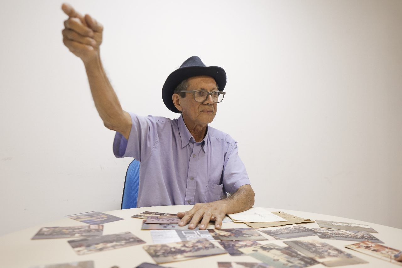 Foto de Severino Vitorino sentado em uma cadeira azul, usando uma camisa de manga curta lilás e um chapéu preto. Ele é um homem branco, idoso, de óculos grandes e está gesticulando com o braço direito levantado e apontando para cima. A mão esquerda está sobre uma mesa redonda, que está coberta com várias fotografias espalhadas. A parede ao fundo é branca e não possui decorações.