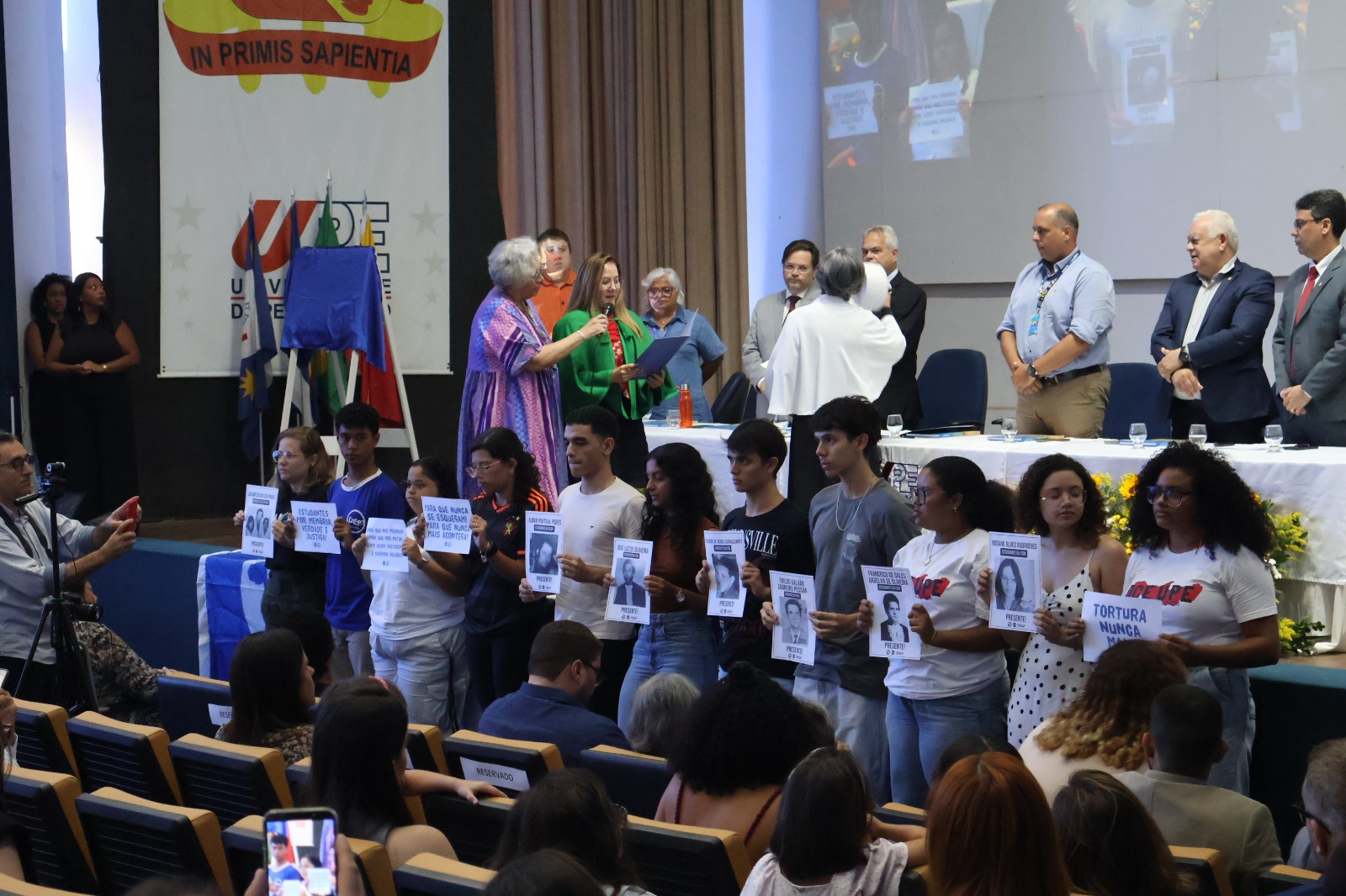 A imagem mostra um evento no auditório da UPE, onde várias pessoas estão participando. Na frente do auditório, há uma fila de pessoas segurando cartazes com fotos e textos. Os cartazes parecem ser de protesto ou homenagem, com algumas mensagens como TORTURA NUNCA MAIS. Atrás dessas pessoas, há uma mesa com várias outras pessoas em pé, algumas segurando documentos ou certificados. No fundo, há uma grande faixa com o texto IN PRIMIS SAPIENTIA e o logotipo da universidade. Algumas bandeiras estão ao lado da faixa.