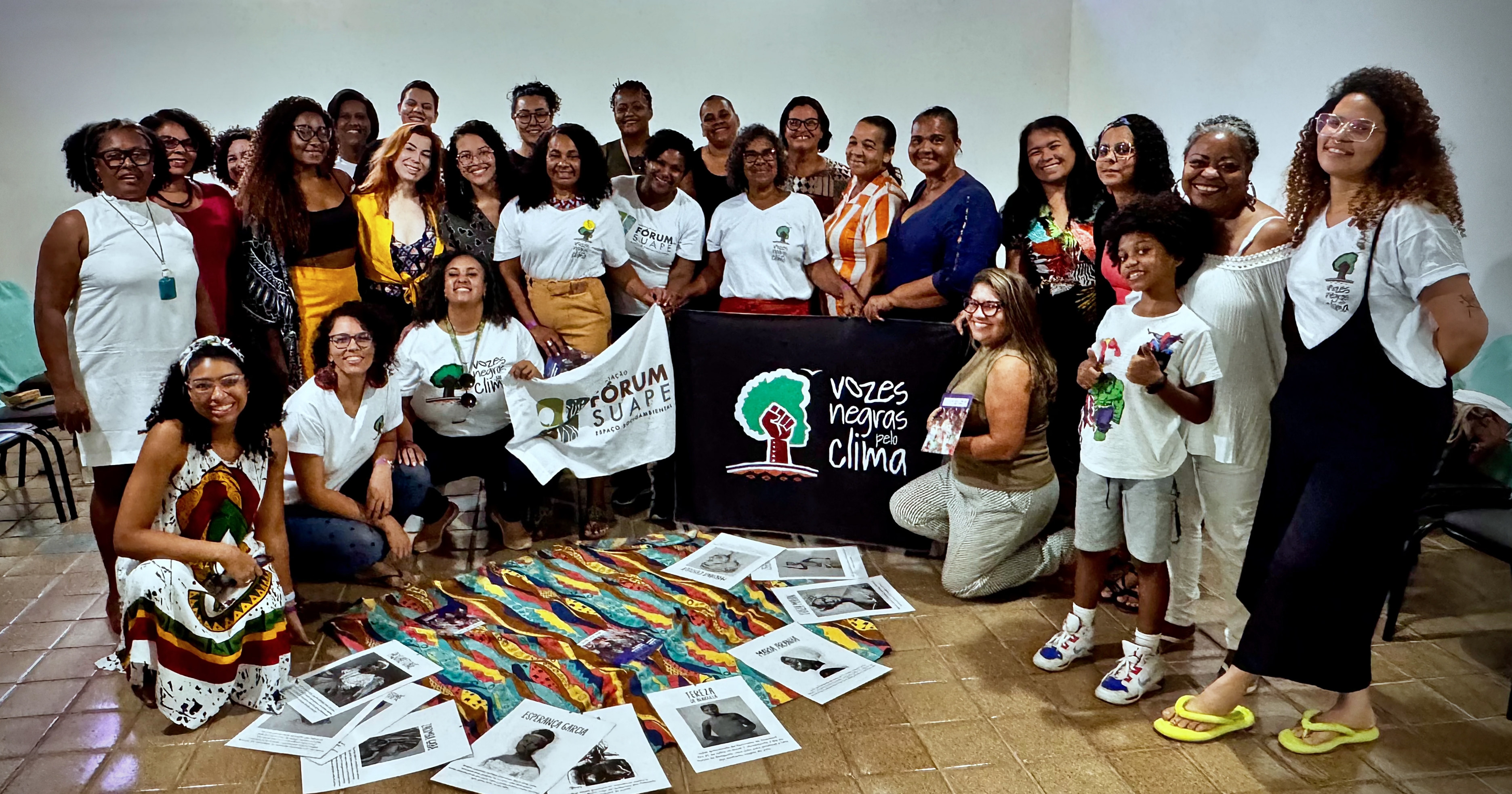 A foto mostra um grupo diverso de pessoas, a maioria mulheres negras, reunidas em uma sala, posando para a câmera com sorrisos. Elas estão em pé e agachadas ao redor de banners que dizem Vozes Negras pelo Clima e Fórum Suape. Há materiais impressos espalhados no chão e algumas pessoas vestem camisetas que parecem relacionadas ao tema de ativismo climático e questões sociais. A imagem transmite um espírito de união e compromisso com causas ambientais e sociais.