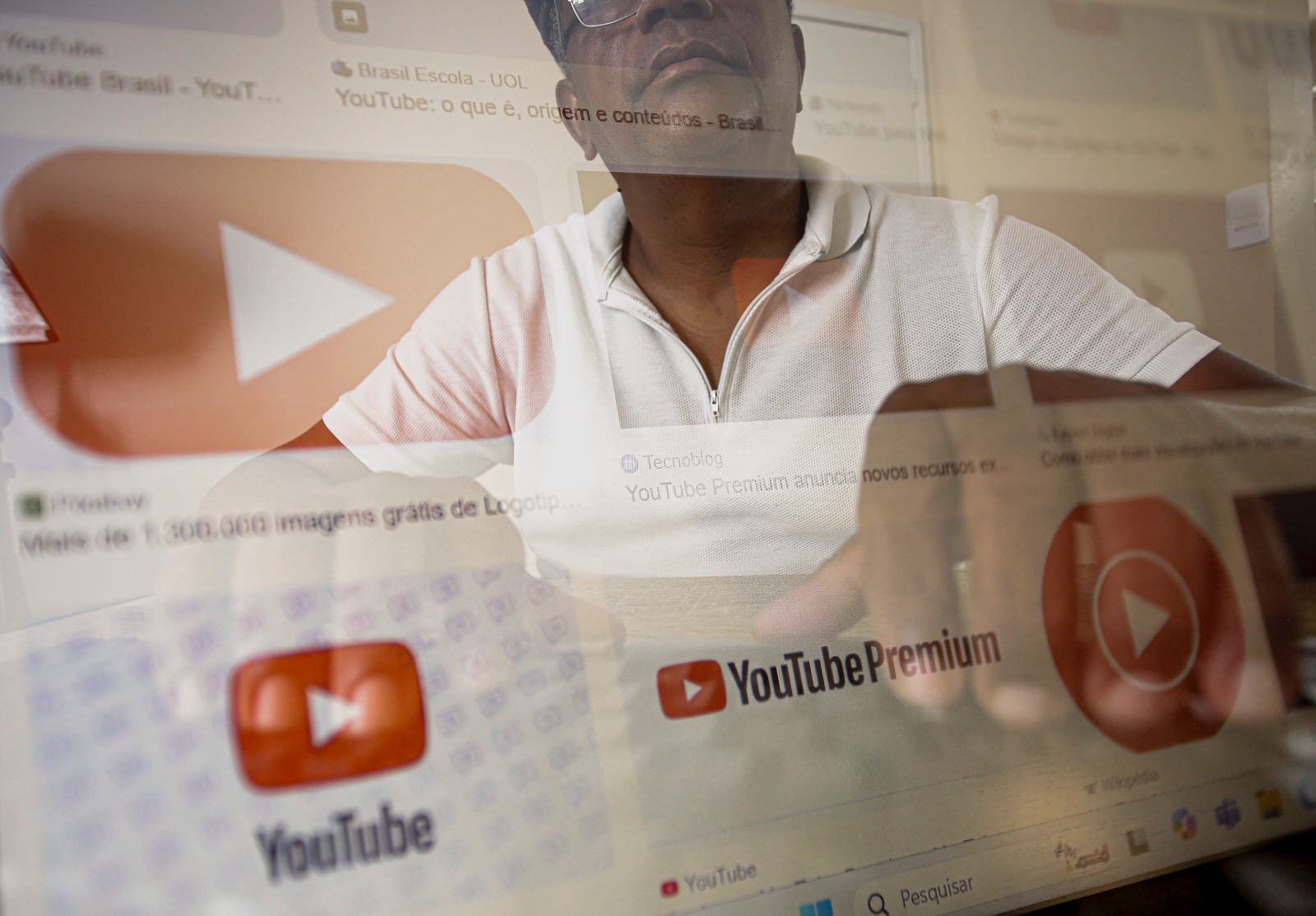 A imagem mostra um homem negro vestindo uma camisa branca com zíper, sentada em frente a uma tela de computador. Na tela, há várias imagens e textos relacionados ao YouTube, incluindo o logotipo do YouTube e o logotipo do YouTube Premium. Alguns dos textos visíveis na tela incluem YouTube: o que é, origem e YouTube Premium anuncia novos recursos. A imagem parece ser uma sobreposição da pessoa e da tela do computador, criando um efeito de transparência.