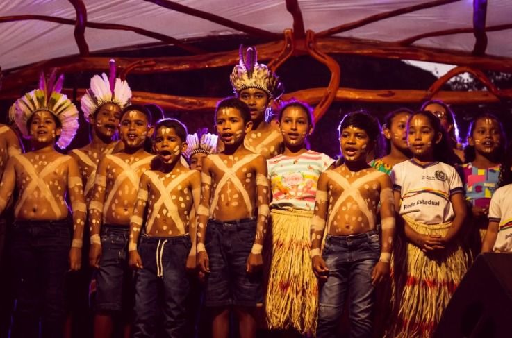 A imagem mostra um grupo de crianças realizando uma apresentação cultural. Elas estão enfileiradas, algumas vestidas com trajes tradicionais, incluindo pinturas corporais em formato de “X” branco no peito e braços, e algumas usando saias de palha e cocares indígenas. A expressão das crianças é de seriedade e concentração, como se estivessem cantando ou recitando algo. A iluminação é amarelada, dando um tom quente à cena, enquanto o cenário de fundo é decorado com elementos naturais, evocando um ambiente ligado à cultura indígena.