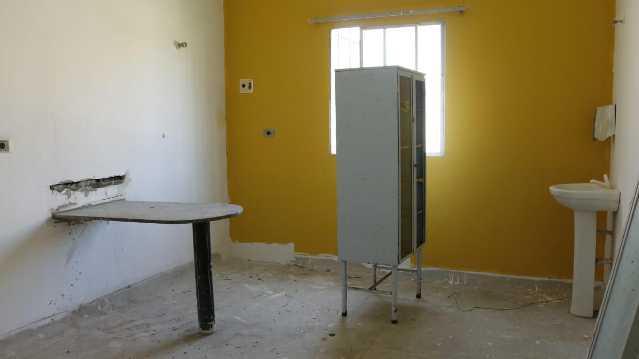 A imagem mostra uma das salas do Hospital Municipal Alzira Figueiredo, em Itamaracá. É um ambiente interno simples e um pouco desgastado. A parede à direita está pintada de amarelo, enquanto a da esquerda é branca e apresenta sinais de desgaste, incluindo um buraco onde uma mesa redonda está fixada na parede. A mesa possui uma perna preta e parece ser improvisada. No centro do cômodo, há um armário metálico alto e desgastado, com portas fechadas. À direita, próximo à parede amarela, há uma pia branca com um pequeno espelho acima dela. O piso é de concreto, também com marcas de desgaste, e há alguns fios no chão.