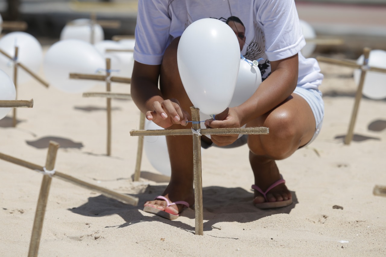 Imagem mostra uma pessoa abaixada, com camisa branca, amarrando um balão branco em uma cruz de madeira fixada na areia da praia de Boa Viagem