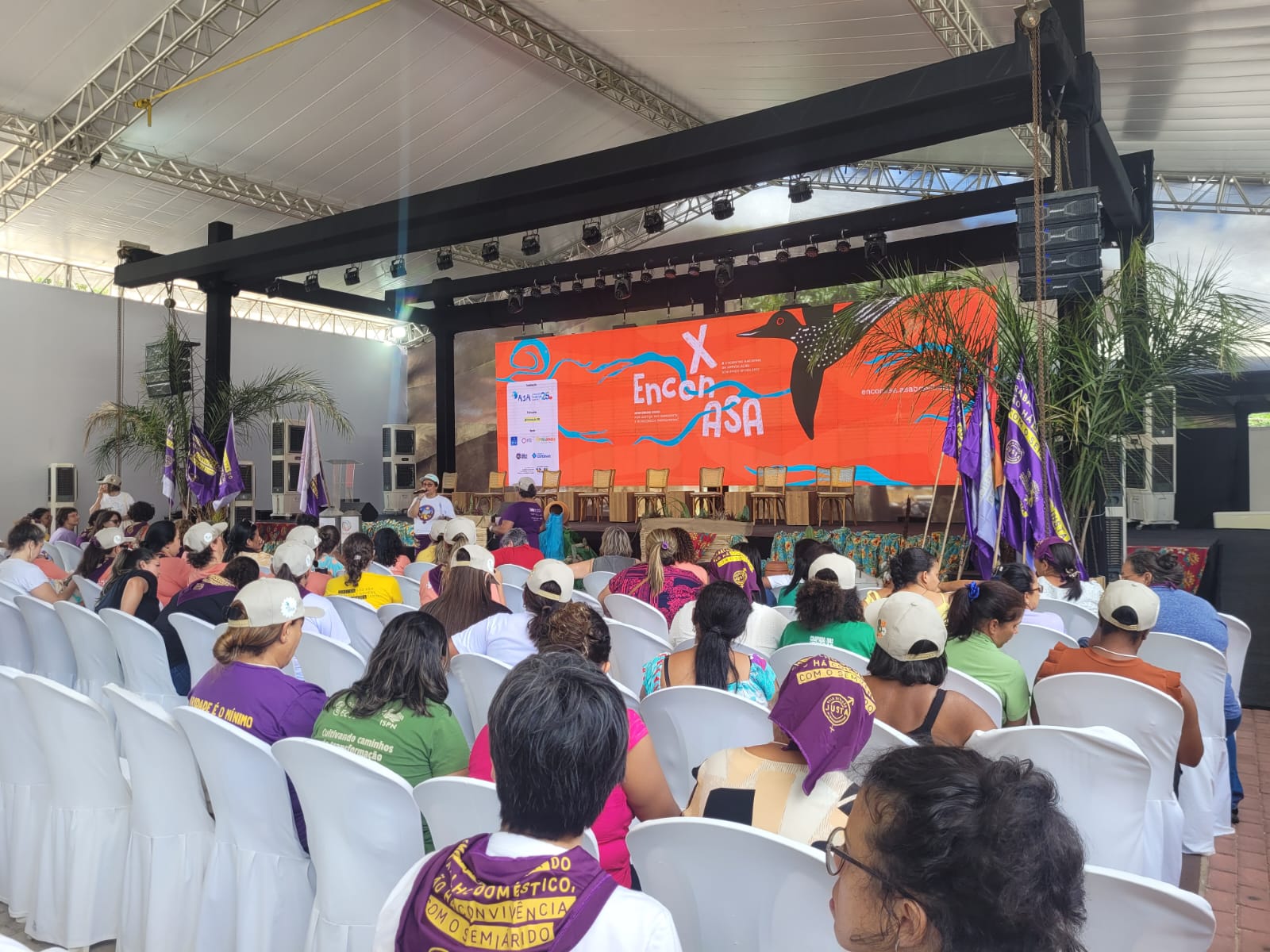 A imagem mostra um evento em um local coberto com um palco ao fundo exibindo X Encontro ASA em uma tela colorida. O público está sentado em cadeiras brancas, e algumas pessoas usam bonés e camisetas com mensagens. Há bandeiras roxas e decoração com plantas tropicais ao redor do palco.