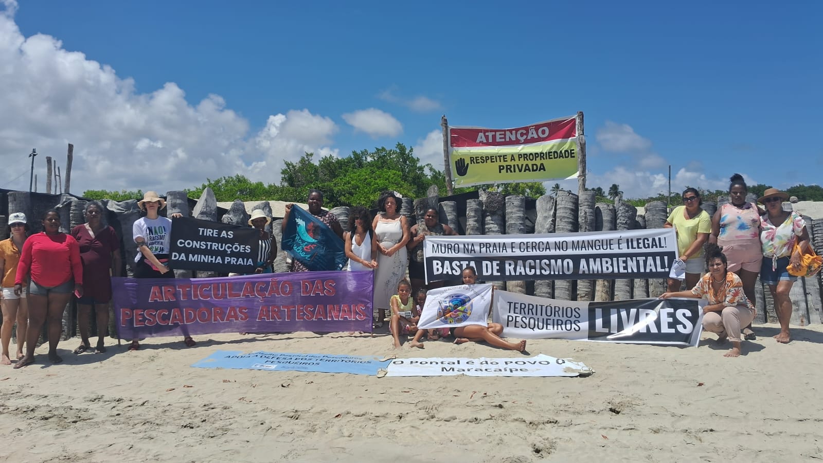 A foto mostra um grupo de pessoas em uma praia segurando faixas e cartazes de protesto. As mensagens incluem frases como Tire as construções da minha praia, Muro na praia e cerca no mangue é ilegal! Basta de racismo ambiental, e Territórios pesqueiros livres. Ao fundo, há uma cerca de madeira e vegetação, destacando o contraste entre a área natural e as reivindicações do grupo.