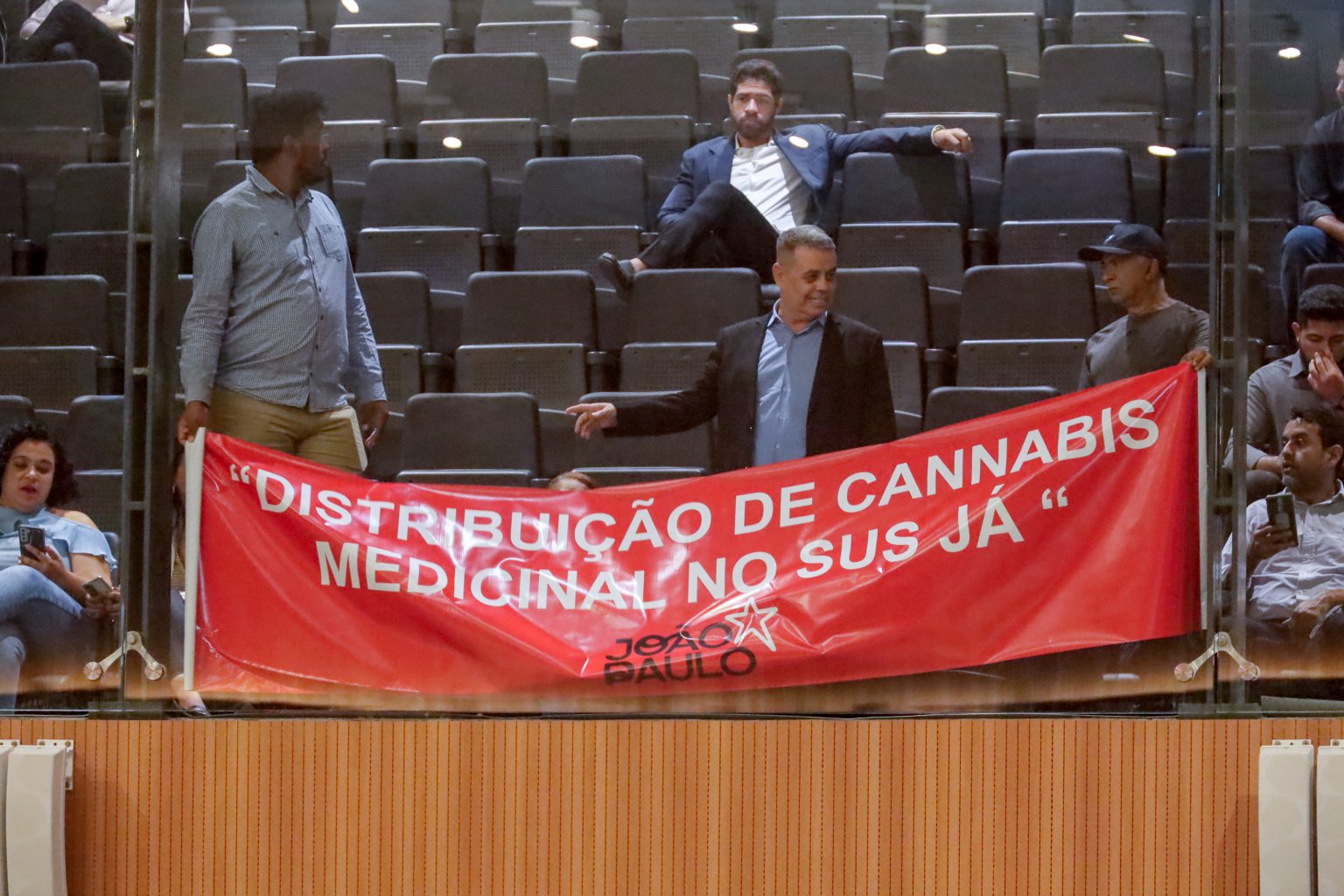 A imagem mostra um grupo de pessoas em um auditório, segurando uma faixa vermelha com a seguinte mensagem em letras brancas: DISTRIBUIÇÃO DE CANNABIS MEDICINAL NO SUS JÁ. A faixa também contém o logotipo do João Paulo com uma estrela. As pessoas estão em pé ou sentadas nas cadeiras do auditório, que estão parcialmente ocupadas.