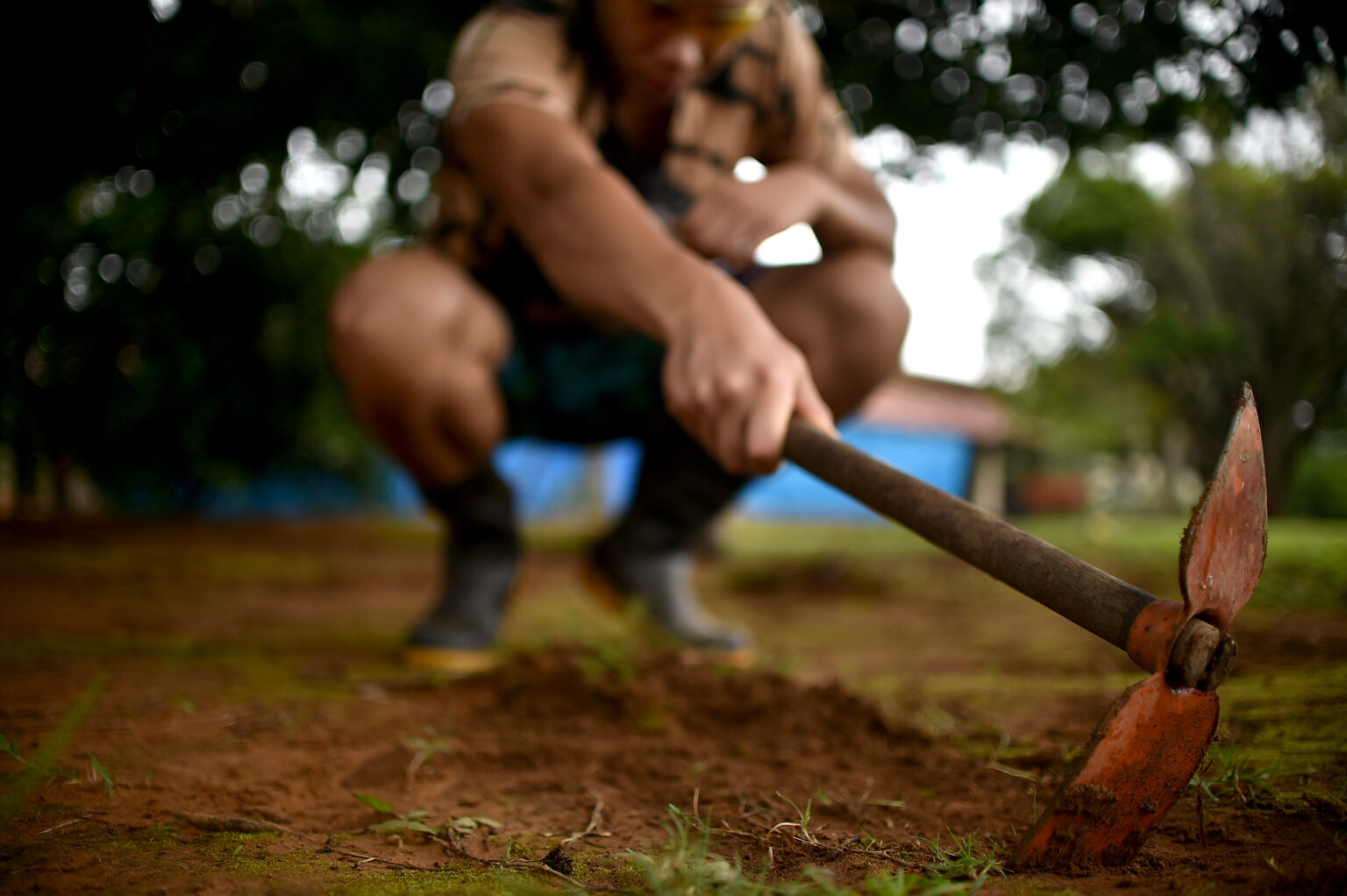 A foto mostra uma pessoa agachada e manuseando uma enxada para trabalhar a terra. O foco está na ferramenta e no solo, enquanto a figura da pessoa aparece desfocada ao fundo.