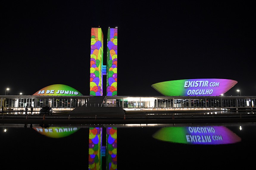 A foto mostra o Congresso Nacional do Brasil à noite, iluminado com projeções coloridas. A cúpula à esquerda exibe a data 28 DE JUNHO e a cúpula à direita apresenta a frase EXISTIR COM ORGULHO, em apoio à causa LGBTQIA+. O reflexo da iluminação é visível na água em frente ao prédio.