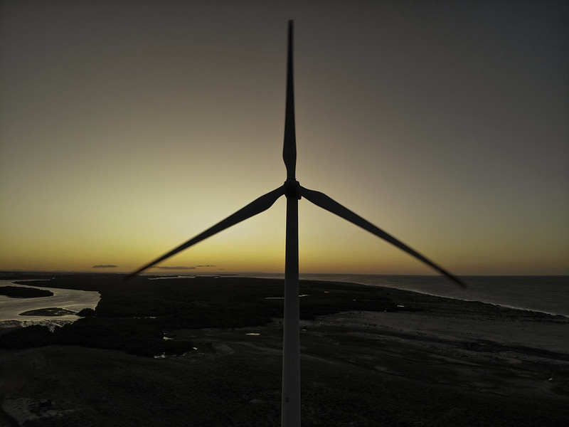 A imagem mostra um aerogerador em silhueta contra o fundo de um pôr do sol ou amanhecer, com uma paisagem costeira ao fundo que inclui água, vegetação e mar.