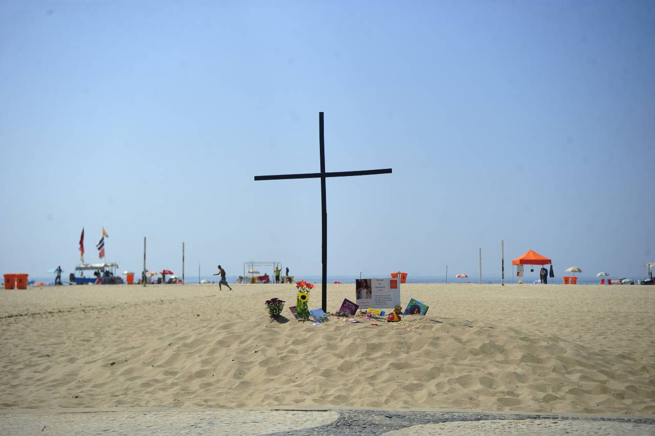 A imagem mostra uma praia de areia clara sob um céu limpo e azul. No centro da imagem, há uma grande cruz preta fincada na areia, que parece ser um memorial improvisado. Ao redor da cruz, há flores e objetos colocados na areia, incluindo girassóis e outros arranjos florais, além de algumas pequenas placas e homenagens com mensagens escritas, provavelmente dedicadas a uma pessoa em memória. Mais ao fundo, algumas pessoas estão na praia, algumas debaixo de guarda-sóis coloridos, enquanto outras estão de pé ou caminhando pela areia