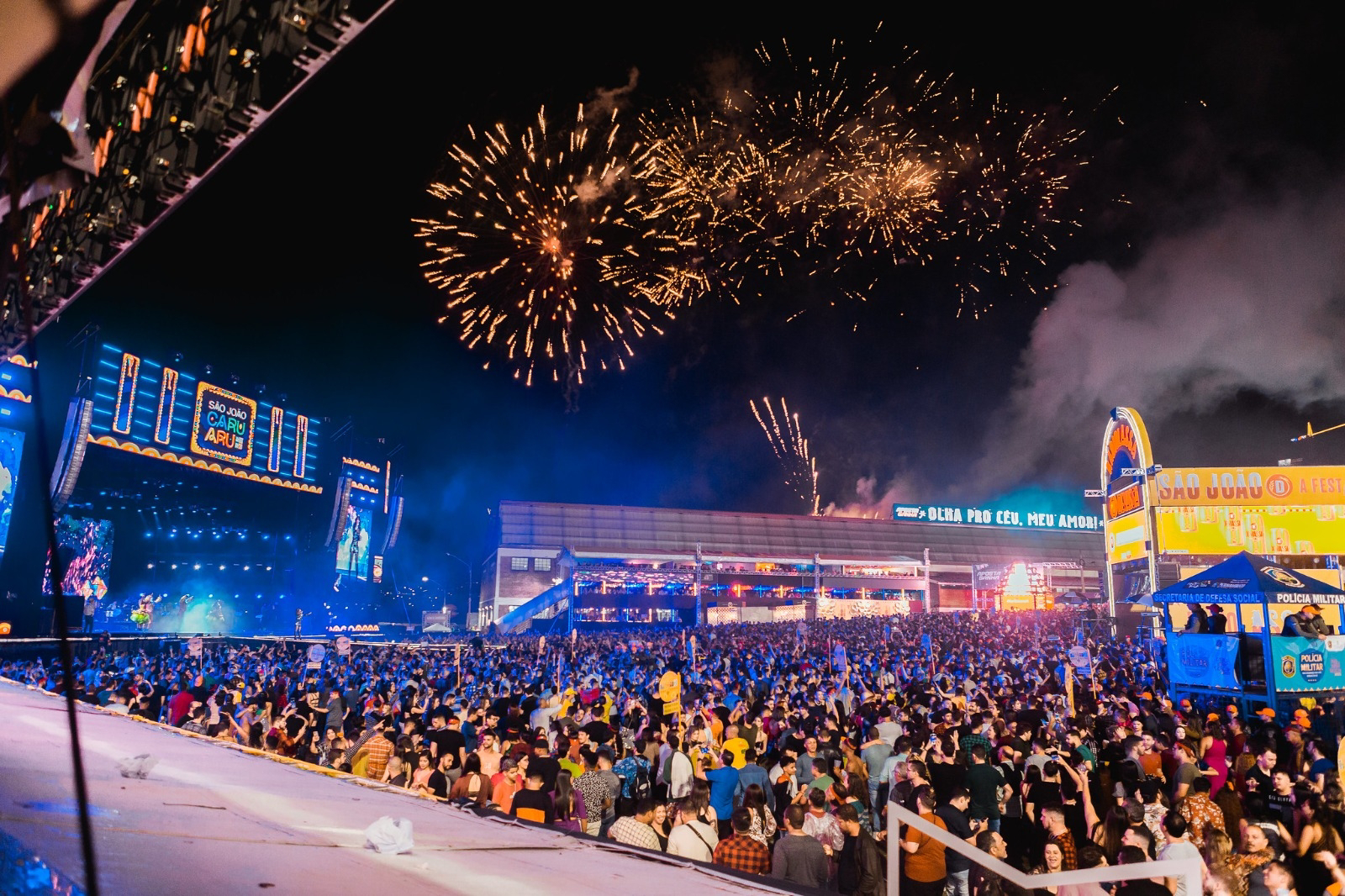 A imagem do link mostra uma cena típica das festividades de São João em Caruaru, Pernambuco. Em primeiro plano, há uma grande área aberta com pessoas celebrando ao redor de barracas decoradas com bandeirinhas coloridas. Ao fundo, observa-se um palco iluminado, onde artistas provavelmente se apresentam. A decoração é vibrante, com muitas luzes e cores tradicionais do São João, como verde, amarelo e vermelho, além das clássicas bandeirinhas que atravessam o espaço