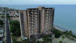 A imagem é a vista aérea de um prédio alto e abandonado à beira-mar. O edifício está em mau estado, com várias janelas e varandas sem vidros ou portas, e as paredes externas cobertas de grafites e pichações. A estrutura de concreto está visivelmente desgastada e danificada. Ao fundo, é possível ver o mar e uma faixa de areia, enquanto à esquerda há uma área residencial com casas e ruas. A vegetação ao redor inclui algumas palmeiras e outras árvores.