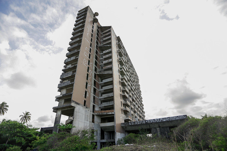 Foto debaixo para cima de um prédio alto e abandonado, com sinais visíveis de deterioração. O edifício tem várias varandas e está coberto de grafites em várias partes, incluindo as paredes externas e a estrutura de concreto na base. A construção parece estar inacabada ou em ruínas, com áreas expostas e sem janelas. Há vegetação ao redor da base do prédio, sugerindo que o local não é mantido. O céu está nublado, e há alguns coqueiros visíveis à esquerda da imagem.