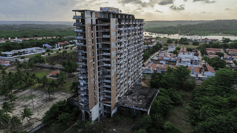 A imagem mostra um prédio alto e abandonado, com várias janelas quebradas e grafites nas paredes externas. O edifício está localizado em uma área com poucas construções e pouco habitada, com várias casas e vegetação ao redor. O prédio está em mau estado de conservação, com sinais de deterioração e abandono. Ao fundo, é possível ver uma paisagem com árvores, um rio e colinas. A imagem foi tirada ao entardecer, com o céu parcialmente nublado.