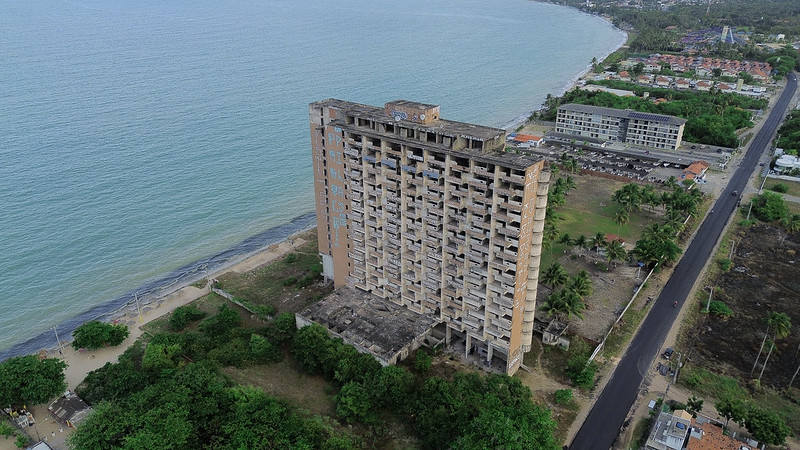 A imagem mostra um prédio alto e abandonado localizado à beira-mar. O edifício tem vários andares e está visivelmente deteriorado, com sinais de abandono e degradação. Ao redor do prédio, há uma área com vegetação e uma estrada pavimentada que passa ao lado. Ao fundo, é possível ver o mar e uma faixa de areia, além de outras construções e áreas urbanas mais distantes. A imagem parece ter sido capturada de uma perspectiva aérea, possivelmente por um drone, oferecendo uma visão ampla da área circundante.