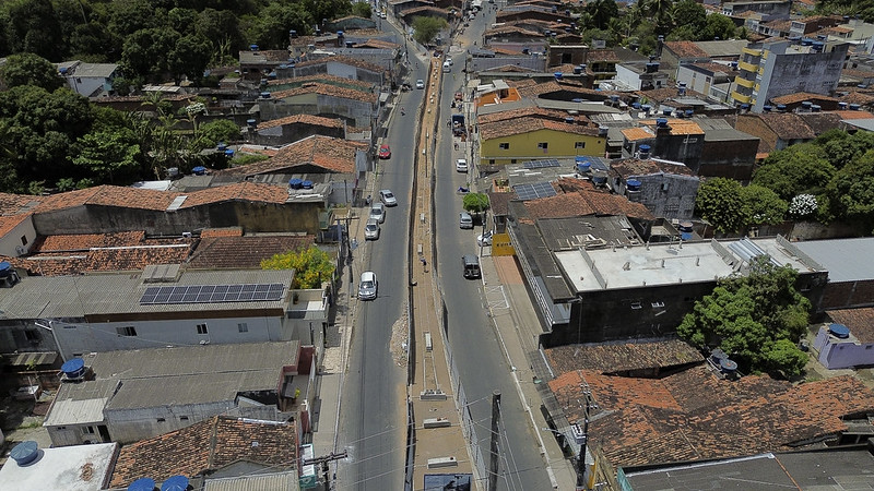 Esta é uma fotografia aérea de uma área urbana, mostrando uma avenida que segue em linha reta no centro da imagem, dividida por um canteiro central estreito. Em ambos os lados da avenida, há casas e construções de tamanhos variados, a maioria com telhados de telhas de barro, característicos de áreas residenciais simples. Algumas casas possuem placas solares no telhado.