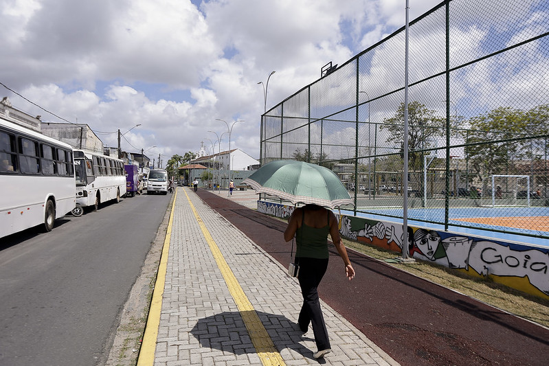 Essa é uma fotografia de um ambiente urbano, tirada durante o dia, sob um céu parcialmente nublado. No lado esquerdo da imagem, há uma rua com ônibus estacionados e outros veículos ao fundo. No centro da foto, uma mulher caminha em uma calçada pavimentada, usando uma sombrinha verde para se proteger do sol. À direita, há uma quadra esportiva cercada por uma grade alta. O muro da quadra possui uma pintura com desenhos artísticos que incluem rostos estilizados e o nome Goiana escrito. O local parece ser um espaço público, possivelmente em uma cidade tropical, dado o clima ensolarado e as roupas leves da mulher.