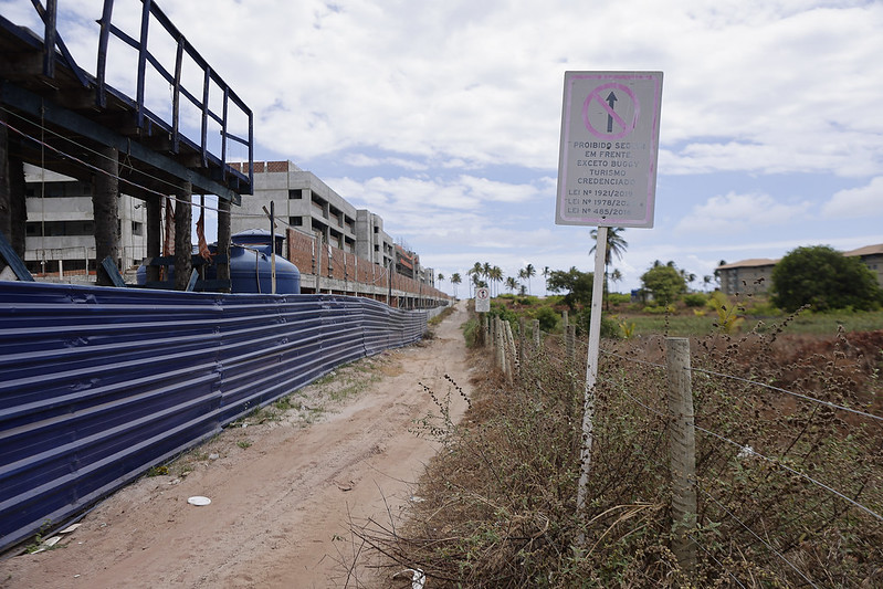 A foto mostra uma visão do nível do solo de uma obra em andamento. Uma cerca de metal azul cobre a frente da construção, que está em fase de acabamento. À direita, há um caminho de terra com uma placa branca avisando sobre proibição de acesso, exceto para moradores credenciados. A placa está desgastada. No fundo, há palmeiras, vegetação e alguns prédios prontos. O céu está parcialmente nublado.