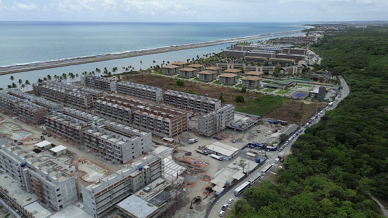 Uma imagem aérea mostra um grande complexo de prédios em construção próximo a uma praia. A área inclui várias estruturas em diferentes estágios de desenvolvimento, com guindastes e equipamentos de construção visíveis. Ao fundo, há o mar, uma faixa de areia e uma paisagem verde ao redor, além de alguns prédios já prontos mais afastados. Uma estrada passa ao lado do canteiro de obras, onde há veículos e outros equipamentos.