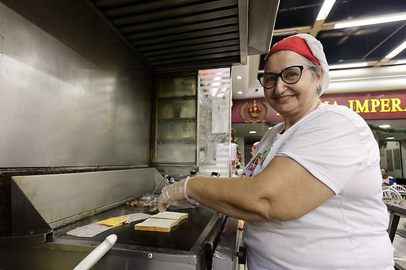 Mulher sorridente vestindo uma touca vermelha e avental branco em frente a uma chapa de cozinha. Ela está preparando sanduíches, com fatias de pão e queijo visíveis na chapa. A cena ocorre dentro da Padaria Imperatriz, identificada ao fundo. A mulher usa óculos e luvas, transmitindo simpatia e cuidado no trabalho.