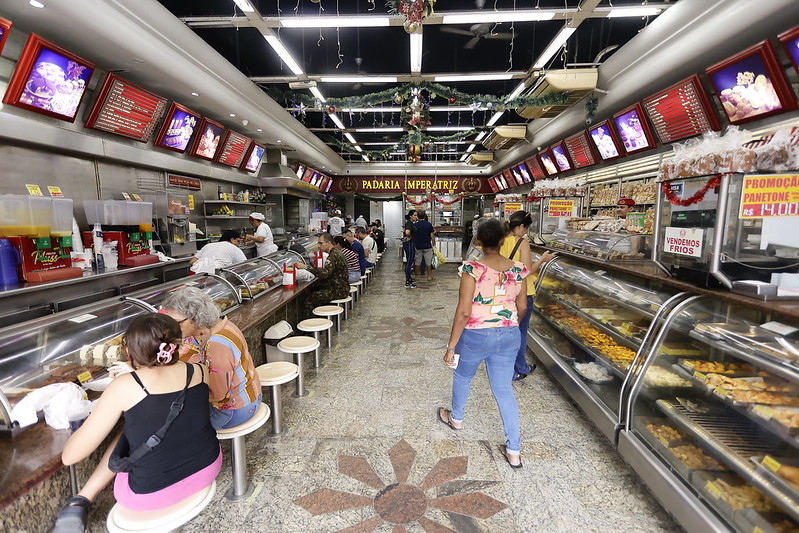 Interior de uma padaria movimentada chamada Padaria Imperatriz. À esquerda, há um balcão com banquetas onde clientes estão sentados comendo. Atrás do balcão, funcionários trabalham preparando alimentos e bebidas. À direita, vitrines exibem pães, salgados e doces. O ambiente é iluminado com luzes de teto fluorescentes, e decorações natalinas pendem do teto.