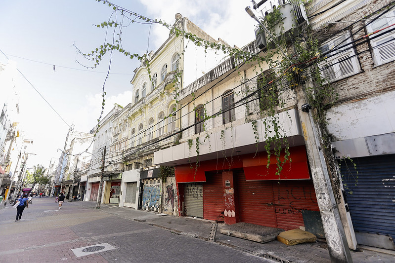 Rua com prédios antigos de fachada histórica, alguns em estado de abandono. Os prédios têm varandas e arcos característicos, mas estão com sinais de desgaste, como vegetação crescendo nas estruturas. As lojas no térreo têm portas fechadas com grafites e sinais de deterioração. Poucas pessoas caminham pela calçada sob um céu claro.