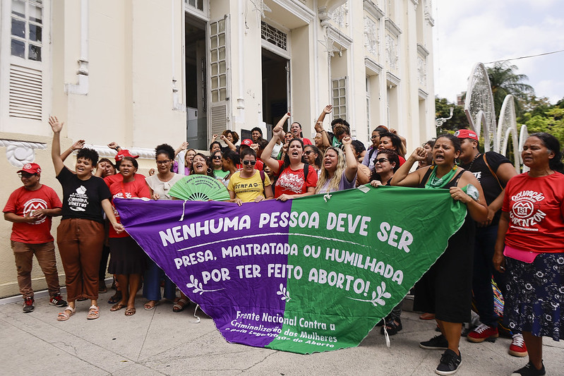 A imagem mostra um grupo de mulheres em frente a um edifício de arquitetura antiga - a Câmara Municipal do Recife - com janelas grandes e abertas. O grupo está segurando uma faixa grande dividade em duas metades nas cores roxa e outra verde, com os seguintes dizeres: NENHUMA PESSOA DEVE SER PRESA, MALTRATADA OU HUMILHADA POR TER FEITO ABORTO! assinada pela Frente Nacional Contra a Criminalização das Mulheres pela Legalização do Aborto