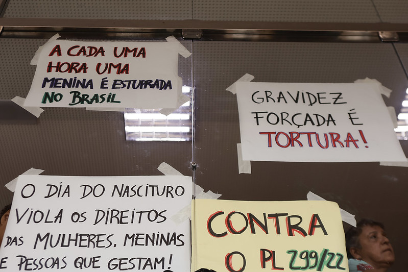 A imagem mostra quatro cartazes colados em uma parede, todos com mensagens em português que parecem ser parte de um protesto ou manifestação. As mensagens dos cartazes são: A cada uma hora uma menina é estuprada no Brasil; Gravidez forçada é tortura! O dia do nascituro viola os direitos das mulheres, meninas e pessoas que gestam! Contra o PL 29/22. Os cartazes criticam a violência sexual e a imposição de gravidez forçada, além de se posicionarem contra um projeto de lei específico (PL 29/22)