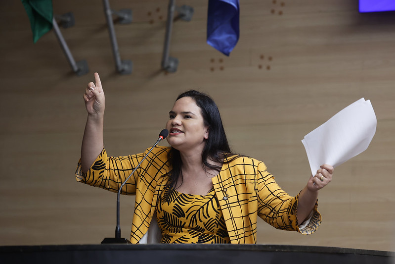 A imagem mostra uma mulher falando em um microfone enquanto segura uma folha de papel. Ela está vestindo um conjunto amarelo com estampa quadriculada e preta. Seu gesto com a mão levantada sugere ênfase ou destaque em sua fala, indicando um ambiente de discurso ou debate público. O fundo mostra elementos de uma sala formal, de um plenário.