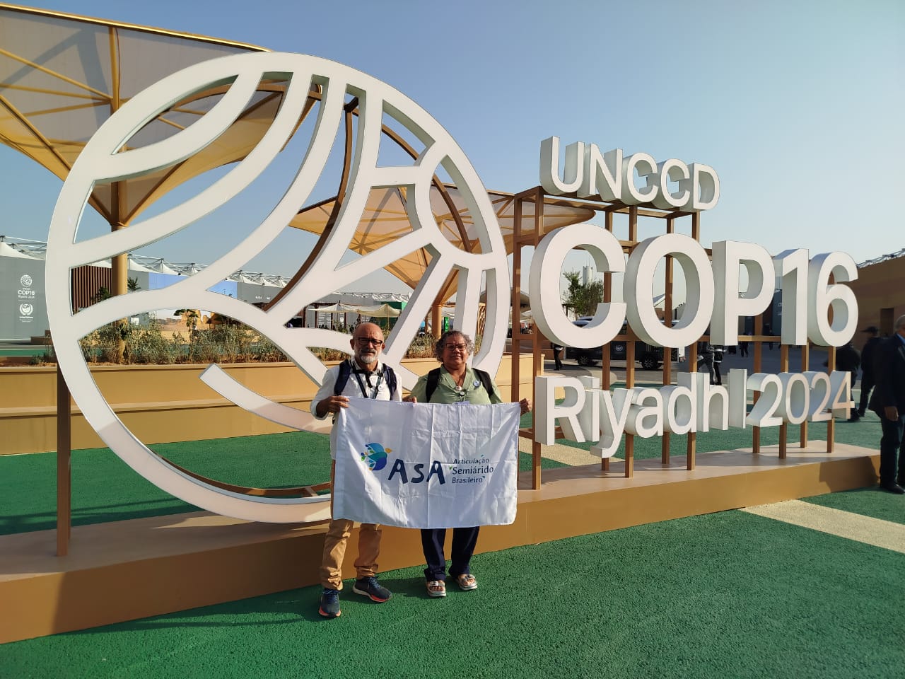 A imagem mostra duas pessoas idosas - um homem calvo, de barba branca e óculos e uma mulher de cabelos grisalhos e óculos - posando juntas em frente a um grande letreiro decorativo que diz UNCCD COP16 Riyadh 2024. O letreiro inclui um design circular estilizado com linhas curvas. As pessoas seguram uma bandeira com o logo e o nome ASA – Articulação no Semiárido Brasileiro. O cenário é ao ar livre, com céu claro e algumas estruturas modernas ao fundo, incluindo tendas e áreas cobertas. O chão é verde, lembrando grama artificial, e há um clima de formalidade, sugerindo que estão participando de um evento internacional sobre questões ambientais ou climáticas.