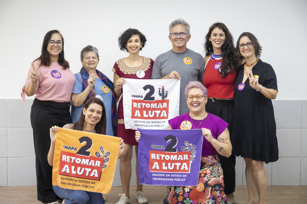 Esta é uma foto de grupo composta por oito pessoas sorridentes posando juntas. São sete mulheres e um homem, Paulo Rubem Santiago. Todas parecem estar em um ambiente interno, como uma sala de reunião, com uma parede clara ao fundo. Algumas delas seguram faixas e bandeiras com mensagens escritas, enquanto outras usam adesivos em suas roupas. As mensagens nas faixas e adesivos dizem: Chapa 2: Retomar a Luta - Adufepe em Defesa da Universidade Pública. As cores das faixas são variadas, incluindo branco, laranja e roxo, com o texto em destaque em vermelho, preto e branco.