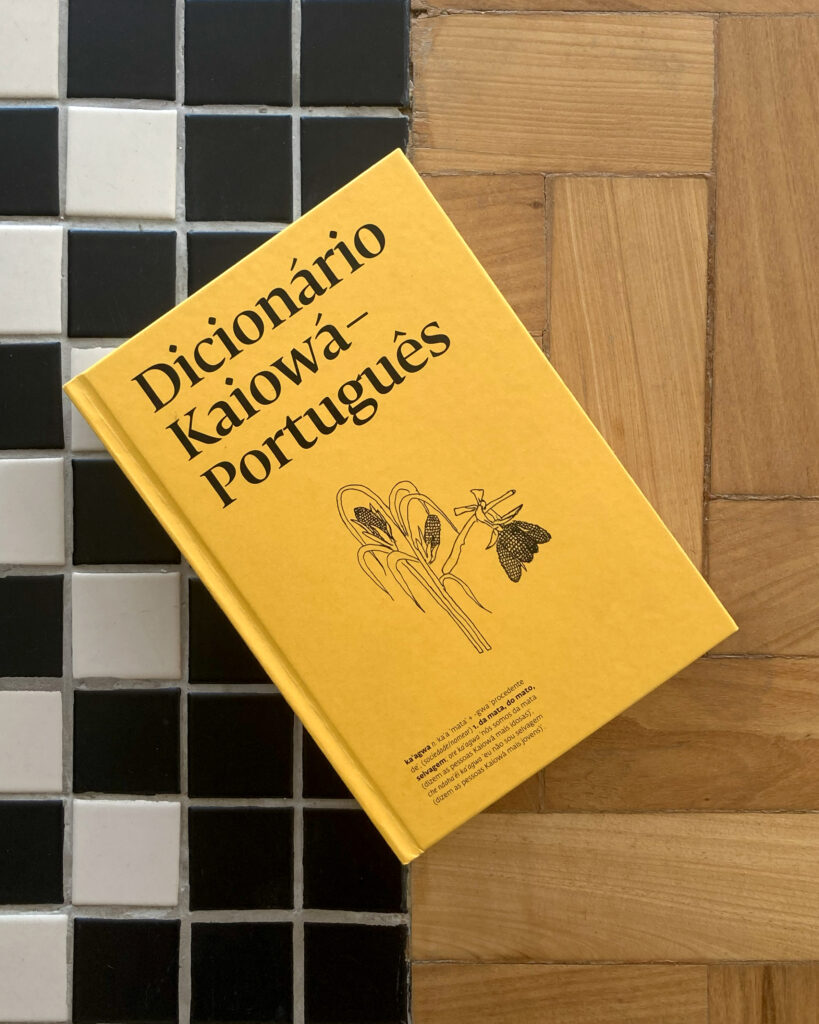 A foto mostra a capa do Dicionário Kaiowá-Português em destaque. O livro tem uma capa amarela com ilustrações simples em preto, incluindo desenhos de uma planta de milho e uma borboleta ou mariposa ao lado. O título está escrito em letras grandes e pretas. O livro está posicionado sobre um chão que combina dois tipos de revestimento: uma parte com azulejos pequenos em preto e branco, formando um padrão quadriculado, e outra parte com madeira clara.