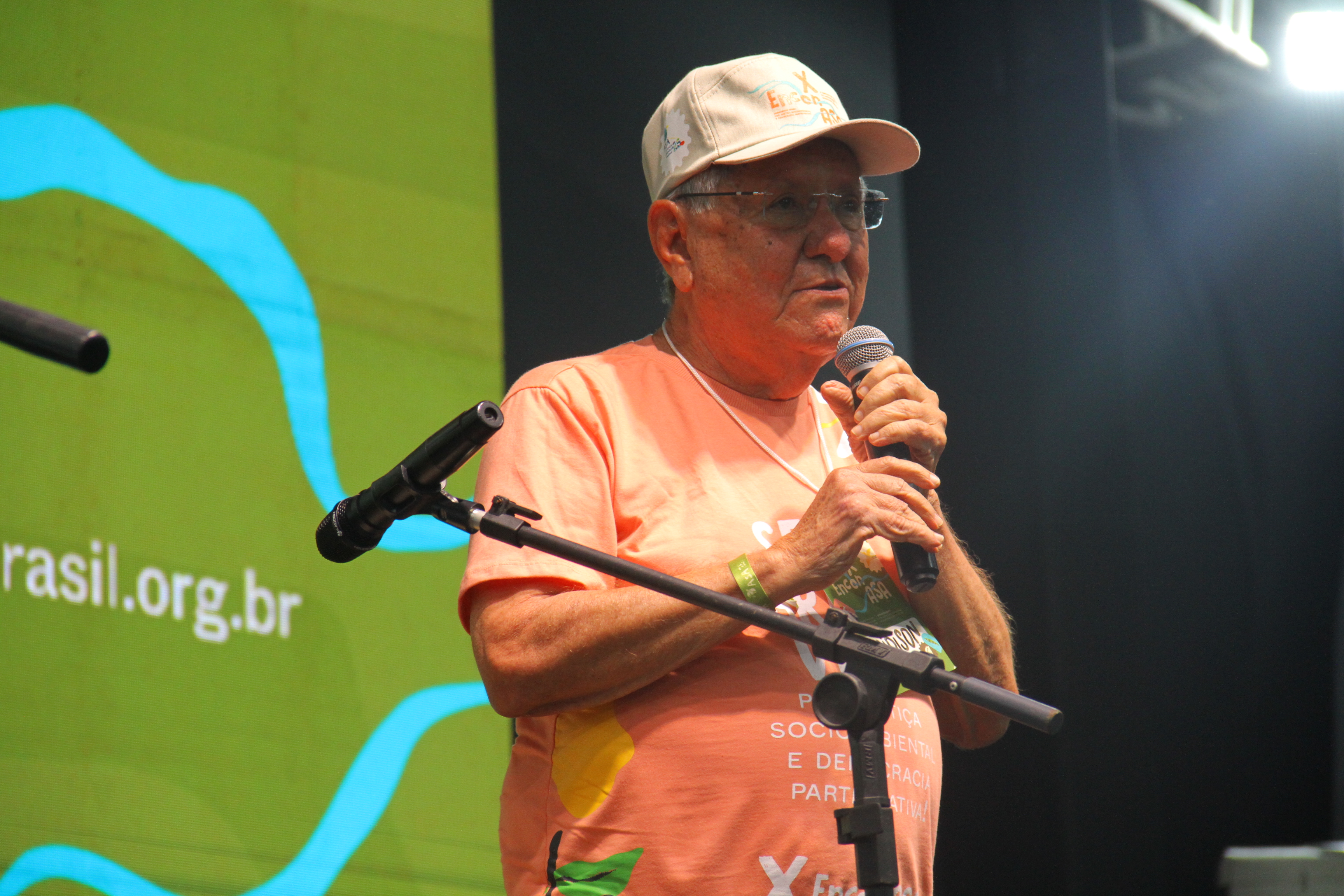 A imagem mostra um homem idoso segurando um microfone enquanto fala em um evento. Ele usa um boné bege com um logo e uma camiseta laranja com texto e gráficos visíveis, embora não completamente legíveis na imagem. Ele está em um palco, com um microfone de pedestal à frente. O fundo tem um banner verde com um logotipo e um endereço de site que inclui brasil.org.br. A iluminação sugere que ele está em um ambiente fechado, com luzes de palco destacando-o. O contexto parece ser uma apresentação ou discurso relacionado a um evento social ou ambiental.