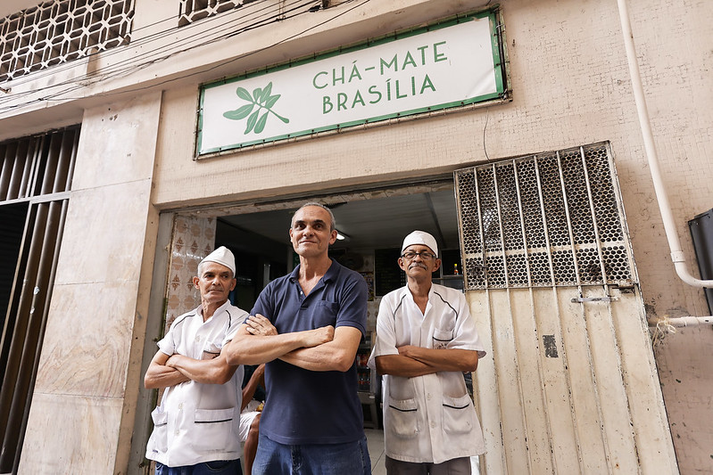 A foto mostra três homens em frente a um estabelecimento chamado Chá-Mate Brasília. O letreiro verde e branco com o nome do lugar aparece em destaque acima da entrada. O prédio é antigo, com paredes desgastadas e um portão de metal ao lado da porta aberta. Os três homens estão de braços cruzados, olhando diretamente para a câmera com expressões confiantes e amigáveis. Eles parecem ser trabalhadores do local. Dois deles, provavelmente funcionários, são idosos e vestem uniformes brancos simples, incluindo toucas na cabeça, como as usadas em lanchonetes ou cozinhas. O terceiro homem, que está ao centro, é alto e magro e veste uma camisa azul escura e calça jeans e pode ser o proprietário ou gerente do lugar. O fundo da foto mostra o interior simples da lanchonete, que parece limpa e organizada.