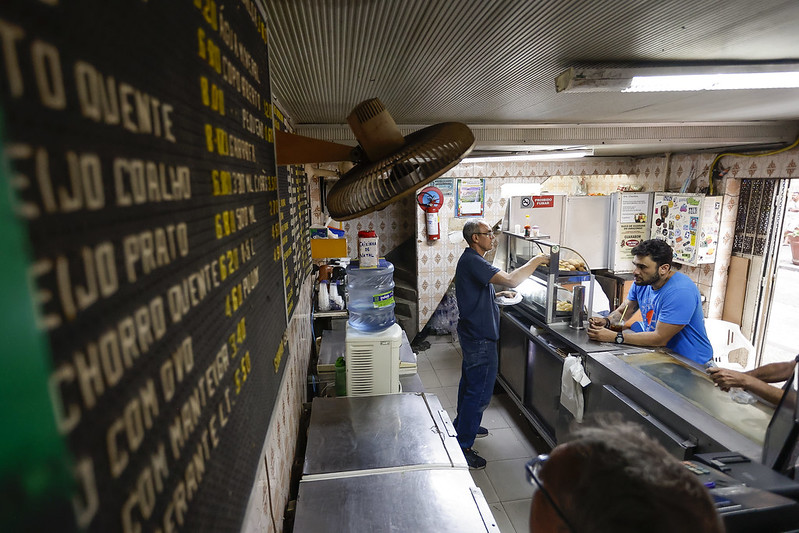 A foto mostra o interior simples de uma lanchonete tradicional. À esquerda, um painel destaca o cardápio com letras amarelas. No centro, um homem alto, magro, de meia idade, José Pinheiro, de camisa azul, está atrás do balcão servindo um cliente, despejando bebida em um copo, possivelmente o famoso chá-mate. O cliente, de camiseta azul, está apoiado no balcão, a espera de ser servido. O balcão de aço inoxidável é organizado, com vitrines exibindo produtos. O ambiente tem paredes com azulejos antigos, uma geladeira com adesivos e um ventilador antigo na parede, reforçando a simplicidade e o clima nostálgico de uma lanchonete acolhedora e tradicional.