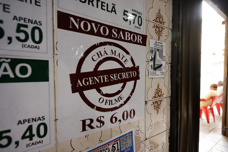 A imagem mostra um cartaz colado em uma parede de azulejos antigos, anunciando um novo sabor de chá-mate chamado Agente Secreto – O Filme, com o preço de R$ 6,00. O cartaz tem um design simples, com fundo branco e letras em marrom. Ao redor, outros anúncios visíveis mostram preços de produtos semelhantes. À direita, a porta aberta revela um ambiente externo, onde duas pessoas estão sentadas em cadeiras plásticas vermelhas