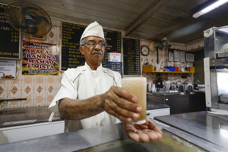 A imagem mostra José Trajano, homem idoso, de óculos, atrás de um balcão, segurando um copo de suco ou vitamina e estendendo-o para a câmera. O homem está vestido com um uniforme branco e um chapéu de cozinheiro. O fundo da imagem revela um ambiente de lanchonete ou bar de sucos, com várias placas e cardápios pendurados na parede. As placas listam diferentes tipos de sucos e vitaminas, como ABACATE, BANANA, MAMÃO e MISTA. Há também um ventilador de teto, um relógio de parede e prateleiras com utensílios e ingredientes. A imagem captura um momento cotidiano em um estabelecimento de alimentação, destacando a interação entre o atendente e o cliente.