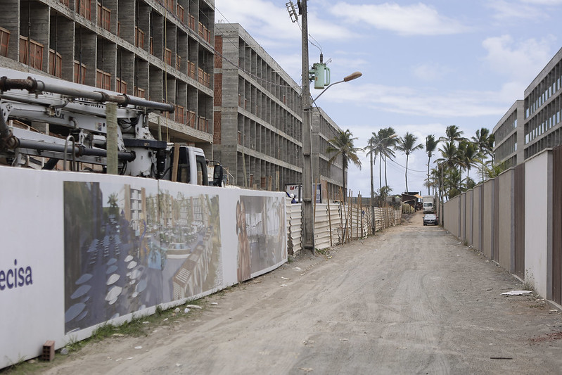 A foto mostra uma rua de terra cercada por muros e prédios em construção de ambos os lados. O muro à esquerda exibe imagens de um projeto arquitetônico de um possível resort, com piscinas e áreas de lazer. Os prédios de concreto estão inacabados, com aberturas para janelas e portas. Ao fundo, há palmeiras altas e um caminhão estacionado. O local parece ser uma área em desenvolvimento, possivelmente turística.