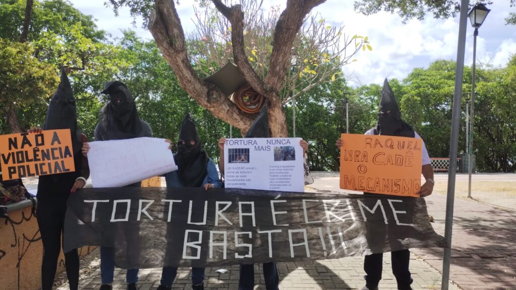 A imagem mostra um grupo de pessoas vestidas com capuzes pretos, segurando cartazes de protesto em um espaço público arborizado. Os cartazes têm mensagens como Tortura é crime basta!, Não à violência policial, Tortura nunca mais e Raquel Lyra, cadê o mecanismo