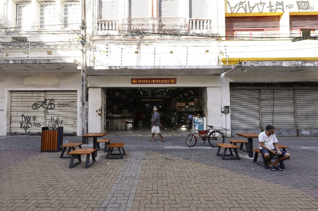 A imagem mostra a fachada da Padaria Imperatriz, com duas mesas e quatro bancos cada em frente. Um homem está sentado, com uma bicicleta estacionada próximo e outro homem passando pelo local. A padaria está entre duas lojas fechadas.